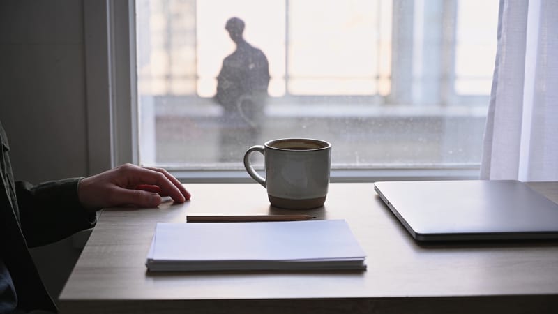 Schreibtisch am Fenster mit Tasse, Bleistift, leerem Papierstapel und zugeklapptem Laptop; eine Hand ruht auf der Tischkante.