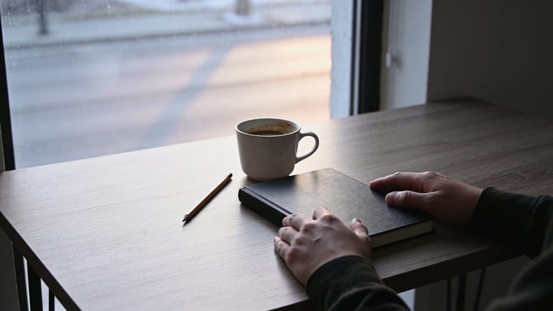 Zwei Hände liegen auf einem geschlossenen schwarzen Notizbuch, daneben eine Tasse Kaffee und ein Bleistift auf einem Tisch am Fenster.