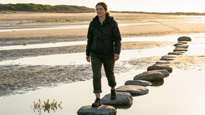 Eine Frau in dunkler Jacke und Wanderhose balanciert auf einer Reihe flacher Steine über nassen Sand bei tiefstehender Abendsonne.
